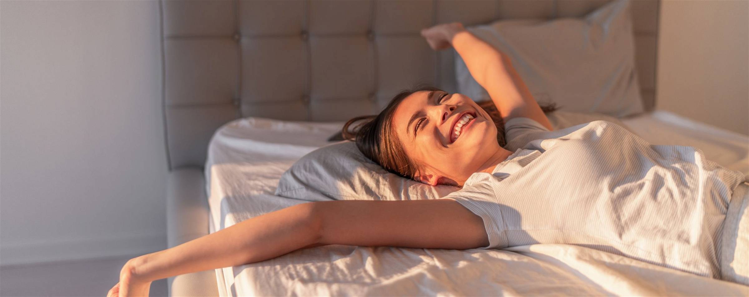 Een uitgeruste gezonde vrouw die zich uitrekt en lachend op bed ligt.